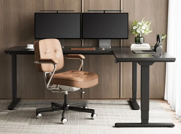 E7L Lite electric standing desk. Black E7L-Lite L-desk with dual monitors, keyboard and flowers, with a tan leather office chair, in a room with wood-panelled walls and sheer curtains.