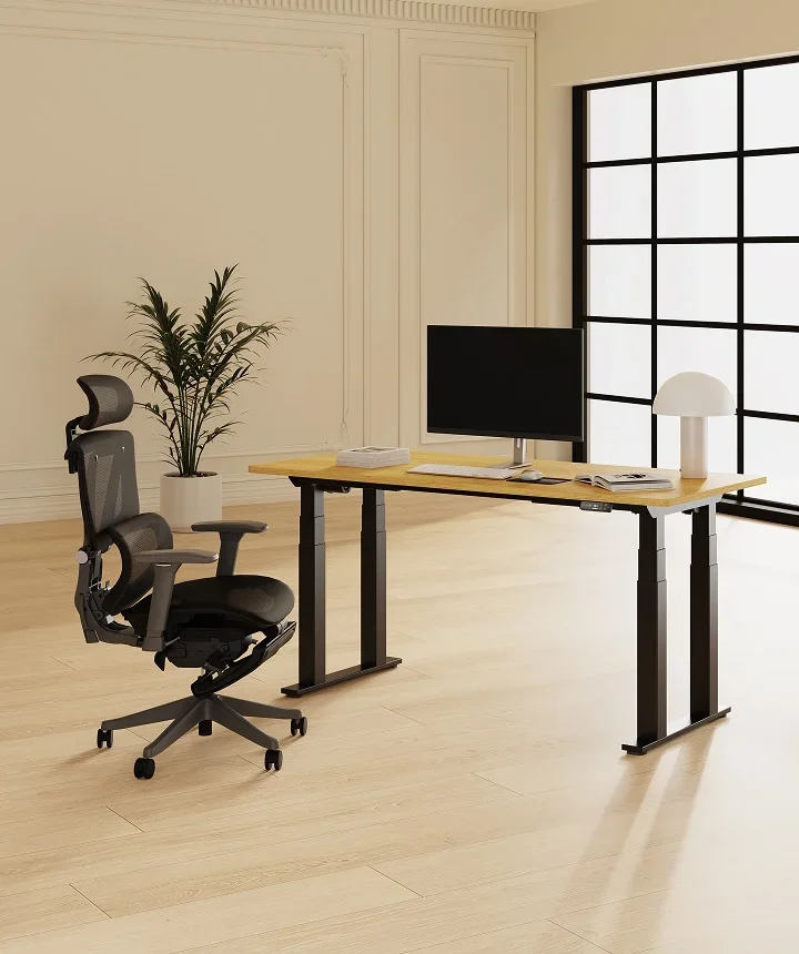 FlexiSpot E7 Plus. E7 Plus with an oak top, monitor and grey mesh chair in a bright room with tall black-framed windows.