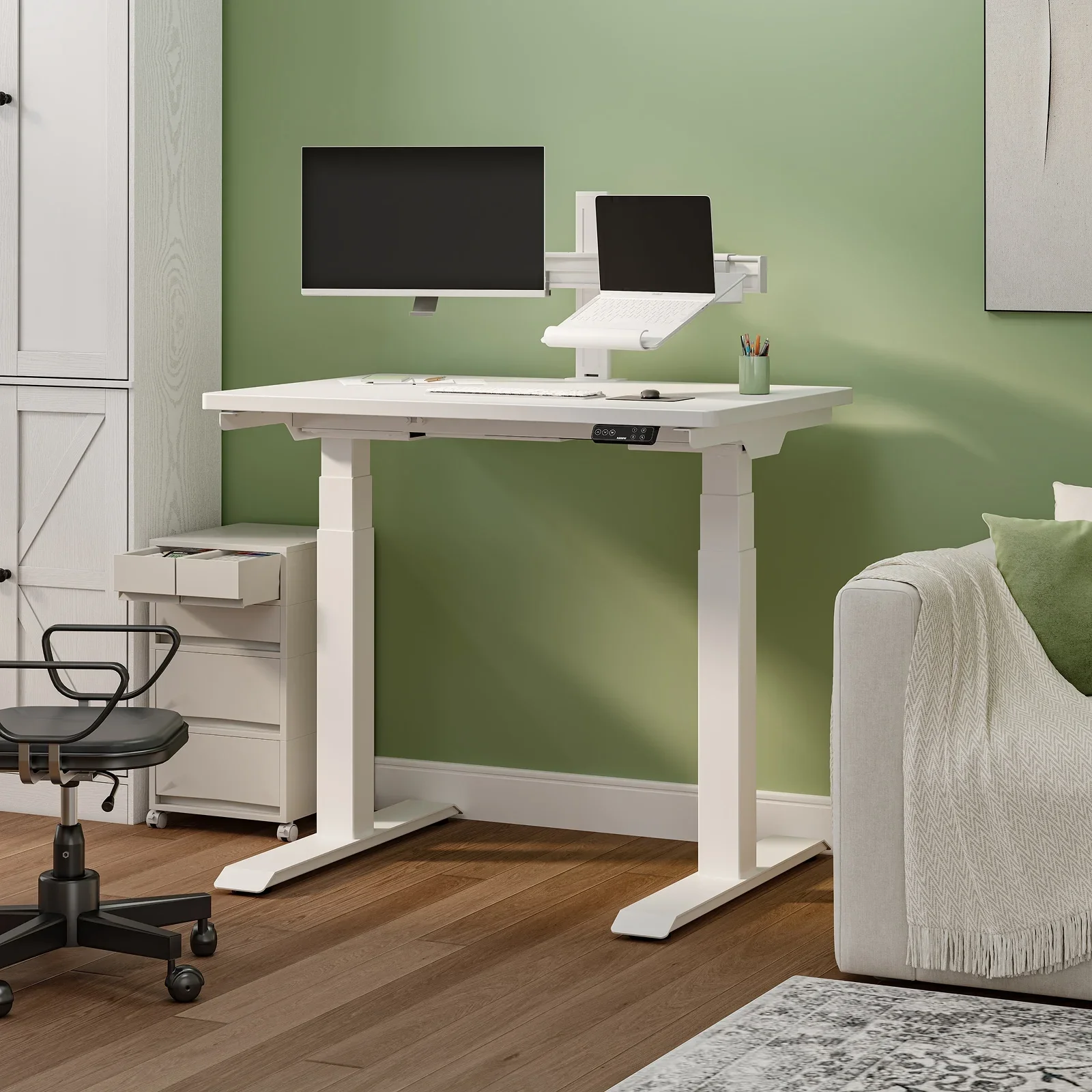 FlexiSpot E7 Mini. White-frame E7 Mini with a white top against a sage green wall, with a monitor on an arm, laptop stand, mobile drawer unit, office chair and sofa.